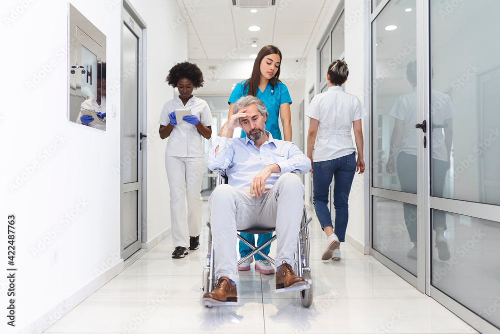 First Floor of the Busy Hospital, Doctors, Nurses and Personnel Busy Working, Assistant Moves Elderly Man in the Wheelchair. New Modern Medical Hospital with Professional Staff.