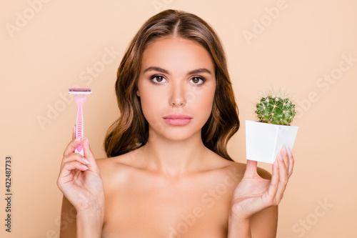 Photo of young serious beautiful girl wear no clothes hold cactus and razor depilation isolated on beige color background photo