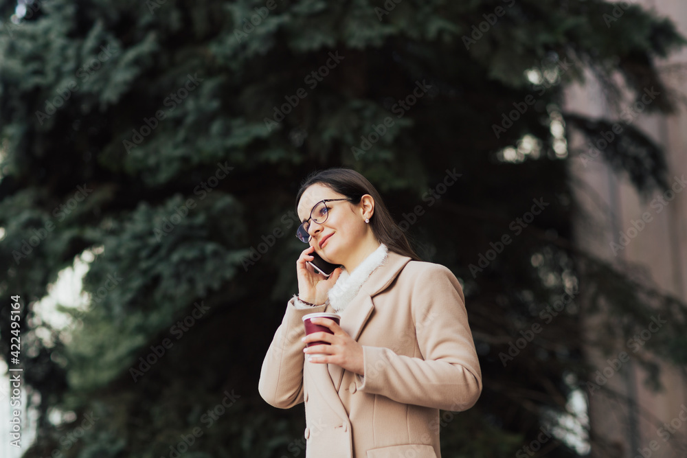 Cheerful young woman wearing a beige coat and eyeglasses talking on her phone near pine and drinking coffee in a paper cup. Cheerful young woman wearing a beige coat and eyeglasses talking on her phone near pine and drinking coffee in a paper cup.