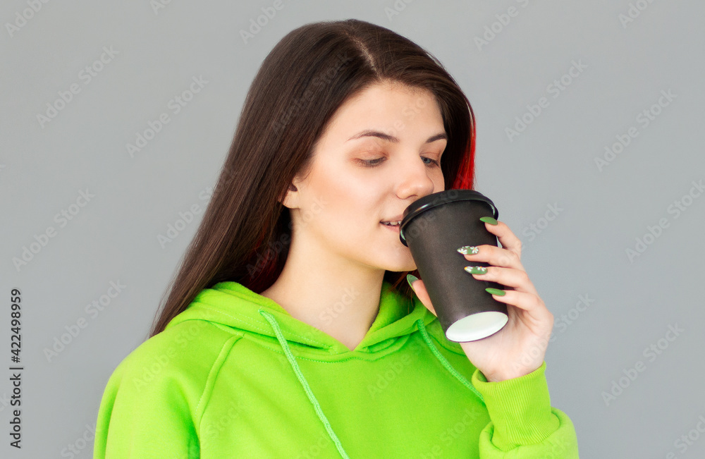 Fototapeta premium Studio shot of girl with holds paper cup of hot coffee, wears green jumper, grey background. Beautiful young woman enjoys aromatic cappuccino.