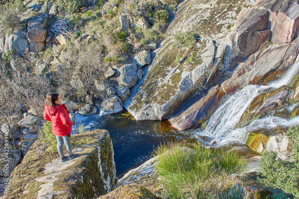 Girl in red jacket perched on a rock watching the waterfall and the natural scenery