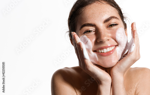 Schilderij op canvas Cheerful female model applies foaming cleanser, has clean fresh healthy skin, smiles broadly, Girl stands bare shoulder against white background