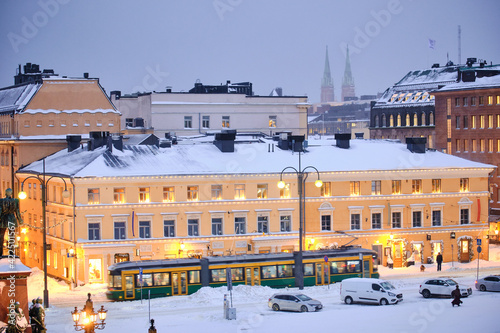 Obraz na płótnie Helsinki, Finland - January 13, 2021: The tram is moving on the Senate Square during the strong snowstorm. The blue building is Helsinki City Museum w salonie