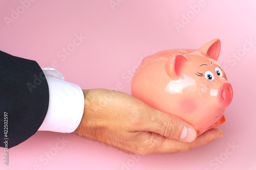 A man holds a piggy bank in his hand on a pink background.