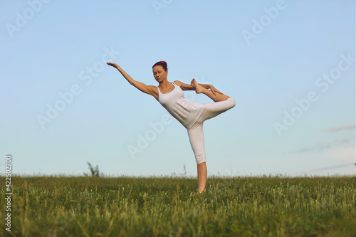 Slim yoga instructor doing Standing Bow pose on green summer meadow under clear blue sky