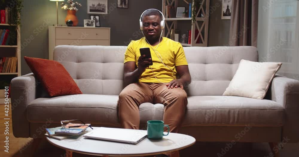 Good-looking African American guy sitting on sofa and texting friend on smartphone at home. Young guy in headphones listening to music, chilling and having fun in evening. Entertainment concept. Good-looking African American guy sitting on sofa and texting friend on smartphone at home. Young guy in headphones listening to music, chilling and having fun in evening. Entertainment concept.