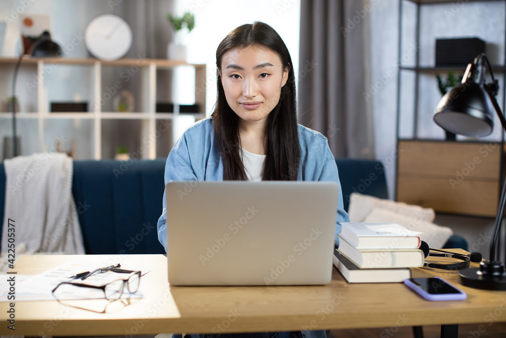 Beautiful asian girl student with long dark hair working indoors at home on wireless laptop while sitting at desk. Female freelancer in denim shirt using computer for remote work. Beautiful asian girl student with long dark hair working indoors at home on wireless laptop while sitting at desk. Female freelancer in denim shirt using computer for remote work.