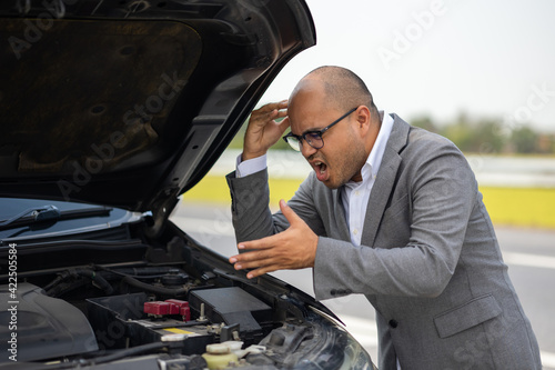 Young asian business has broken down car. He open hood and check the engine. He feeling upset and stressed. Car broken on the side of the road