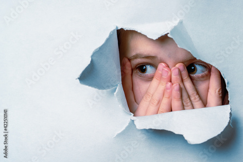 Portrait of young cute scared child girl with wide open eyes looking outside through a hole in the wall. Copy space for text.