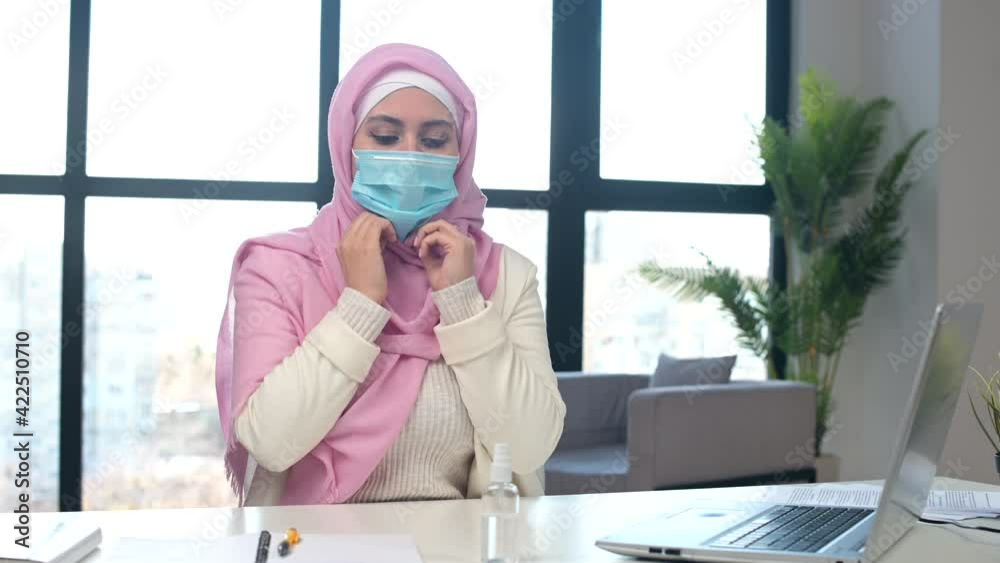 Smiling muslim woman wearing hijab demonstrates how to protect yourself from viral diseases while working in the office. Islamic business woman looks at the camera, puts mask on, sanitizing hands Smiling muslim woman wearing hijab demonstrates how to protect yourself from viral diseases while working in the office. Islamic business woman looks at the camera, puts mask on, sanitizing hands