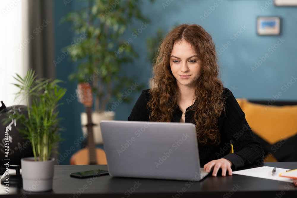 A young student girl is studying remotely from her home. Female uses laptop, browses websites and social media. The girl is sitting at a desk in a cozy living room. A young student girl is studying remotely from her home. Female uses laptop, browses websites and social media. The girl is sitting at a desk in a cozy living room.