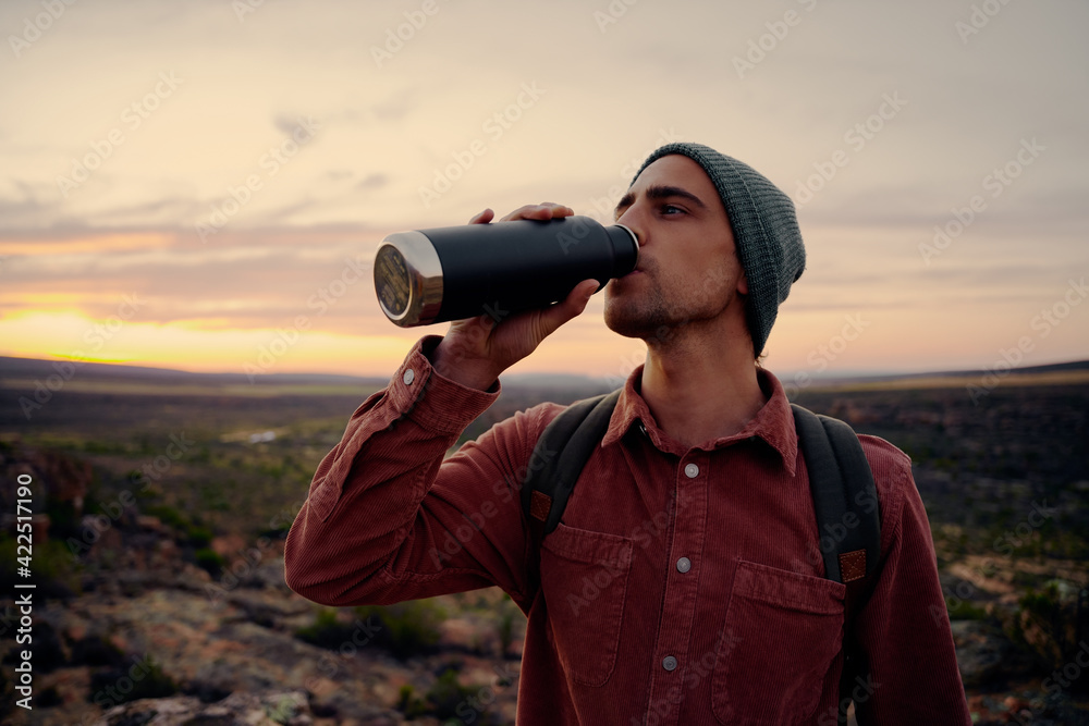 Young male hiker wearing backpack and cap drinking water standing on mountain