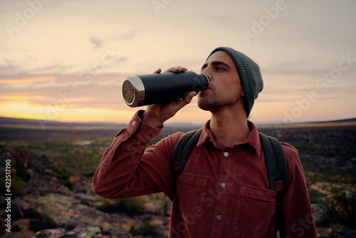 Young male hiker wearing backpack and cap drinking water standing on mountain