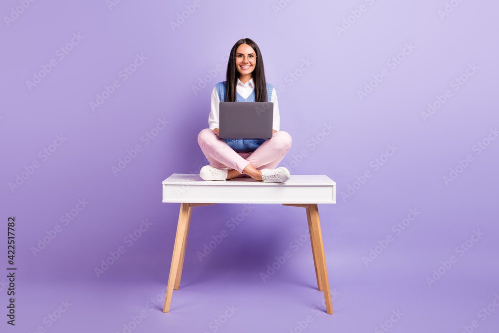 Portrait of beautiful brunette cheerful girl sitting on table in lotus position using laptop studying isolated over purple violet color background Portrait of beautiful brunette cheerful girl sitting on table in lotus position using laptop studying isolated over purple violet color background