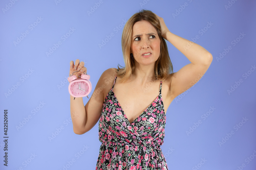 Fototapeta premium Portrait of cheerful young doubtful woman holding alarm clock isolated over purple background