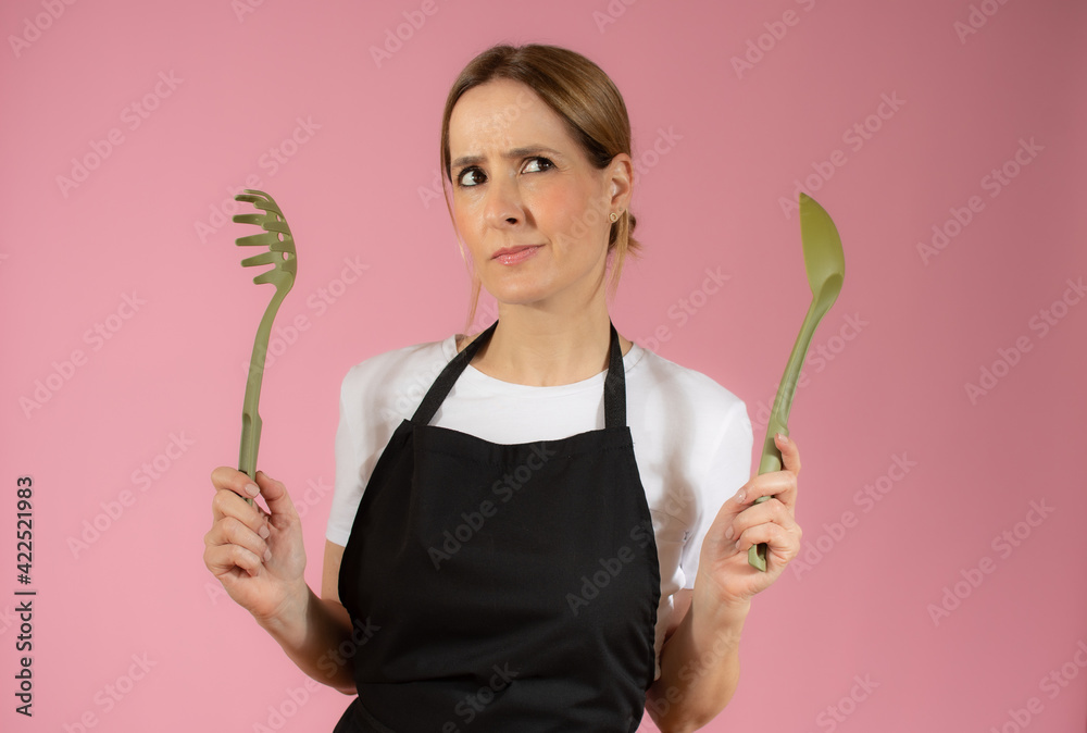 Young caucasian woman holding kitchen utensils against pink background Young caucasian woman holding kitchen utensils against pink background