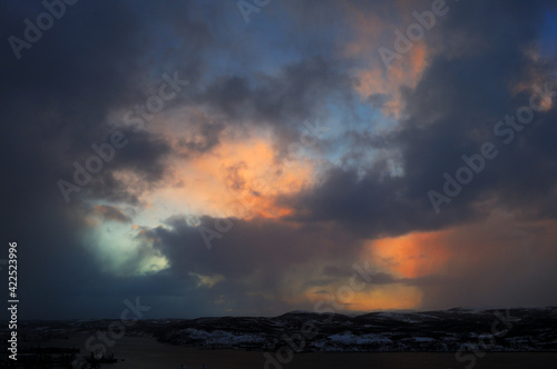 Blue sky with gray clouds over snow-capped mountains. Clouds illuminated by the sun over a dark city and mountains in the snow. Northern nature of the city of Murmansk.