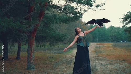 Fantasy woman in black creative dress with raven on hand posing with open arms like bird dreams of freedom flying to sky. Dark fairy with pet. Nature green wild forest pine. Fashion model dark queen