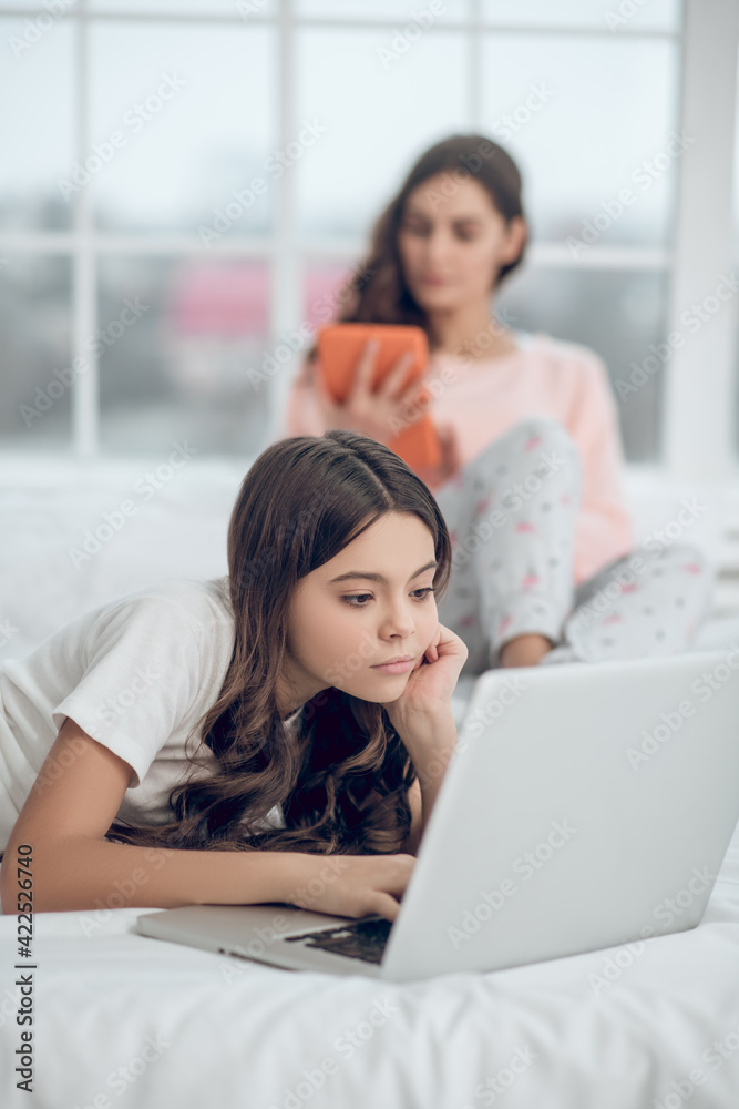 Serious daughter near laptop and mom with tablet Serious daughter near laptop and mom with tablet