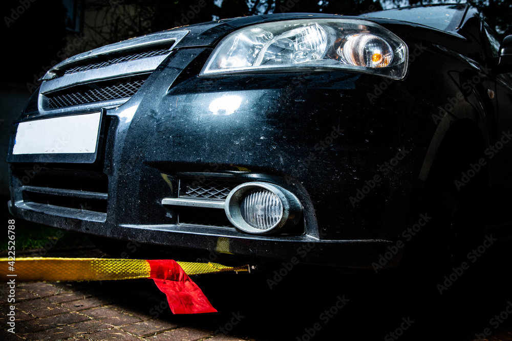 A horizontal view of a black automobile with yellow towline attached to a car towing eyelet. Towing a broken vehicle with a tow rope at night. Car assistance services.
