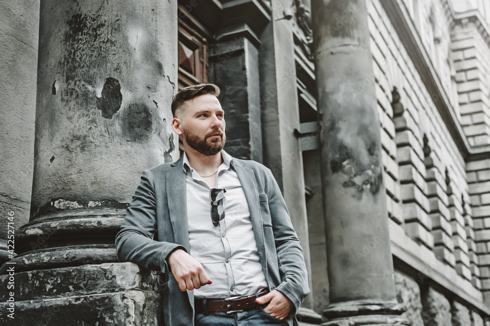Portrait of young successful smart businessman in white shirt, classic suit. Man standing near stone building outdoors. Mobile Office, business concept.