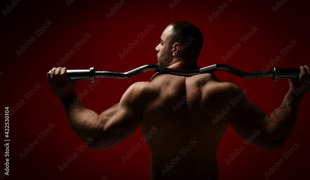 Brutal strong man athlete in stand with his back to camera holding barbell on his shoulders and looks aside over red background. Sport men body concept Brutal strong man athlete in stand with his back to camera holding barbell on his shoulders and looks aside over red background. Sport men body concept