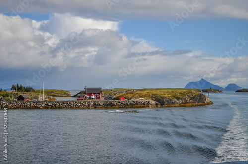 View from ferry to small village with red houses in Helgeland archipelago in the Norwegian sea on sunny summer morning. Boat water trail on water. Donna island silhouette in background