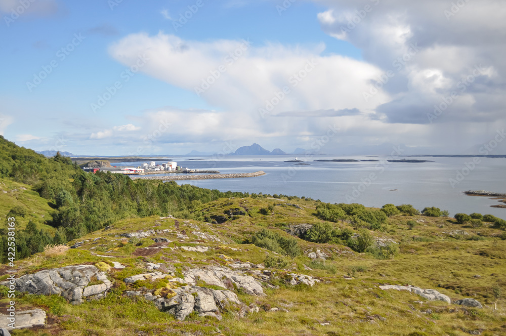 View from Lovund hills to the rocky coastline of Lovundf and Lovund harbor on sunny summer morning. White clouds over the blue sea. Donna island on the horizon.