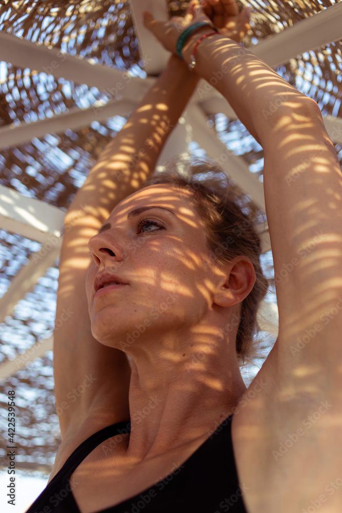 Fototapeta premium Beautiful woman in swimsuit standing under a beach umbrella with contrasting shadows from branches on the face. Summer vacation concept