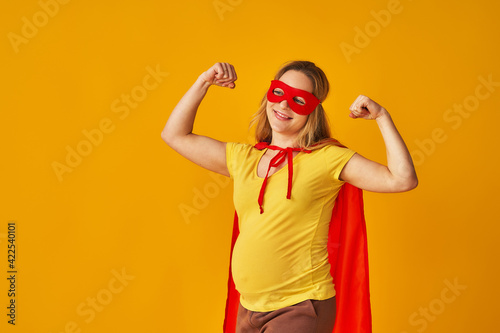 attractive smiling pregnant woman in superhero costume, wearing red mask and cape, showing her strength, muscles, stands on yellow background. concept superpowers girl, feminism, desire to win