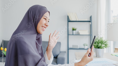 Young Asia muslim businesswoman using smart phone talk to friend by videochat brainstorm online meeting while remotely work from home at living room. Social distancing, quarantine for corona virus.