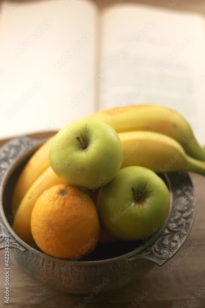 Antique bowl with bananas, apples and lemons and open book on wooden table. Selective focus. Antique bowl with bananas, apples and lemons and open book on wooden table. Selective focus.