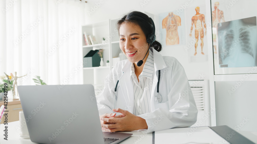 Young Asia lady doctor in white medical uniform with stethoscope using computer laptop talking video conference call with patient at desk in health clinic or hospital. Consulting and therapy concept.