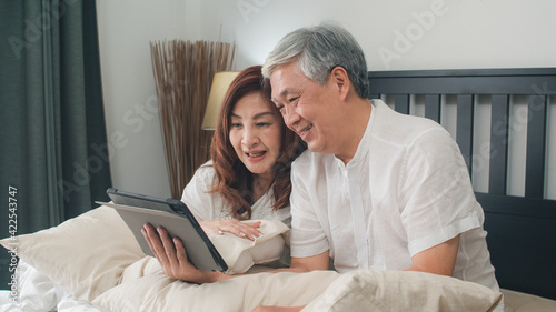 Asian senior couple using tablet at home. Asian Senior Chinese grandparents, video call talking with family grandchild kids while lying on bed in bedroom at home in the morning concept.