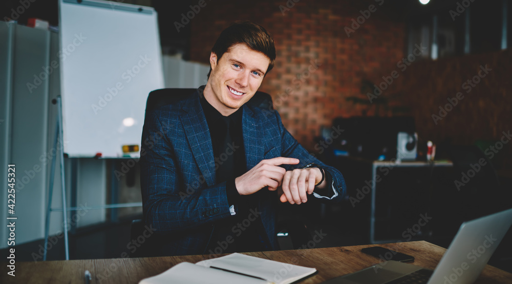 Portrait of cheerful male employer in elegant formal wear smiling at camera while checking clock time, successful Caucasian businessman using modern smartphone during work day in office interior