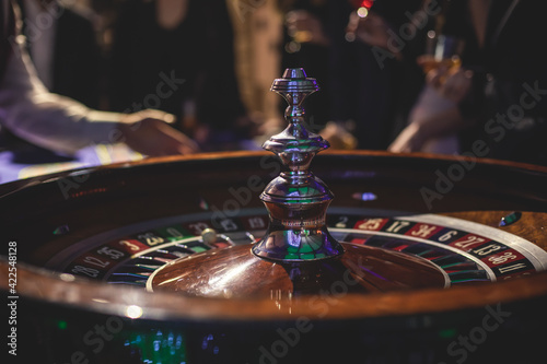 A close-up vibrant image of multicolored casino table with roulette in motion, with casino chips. the hand of croupier, money and a group of gambling rich wealthy people playing in the background