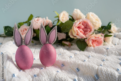 Two pink Easter eggs with bunny ears close-up on a white knitted blanket and a blue background with flowers