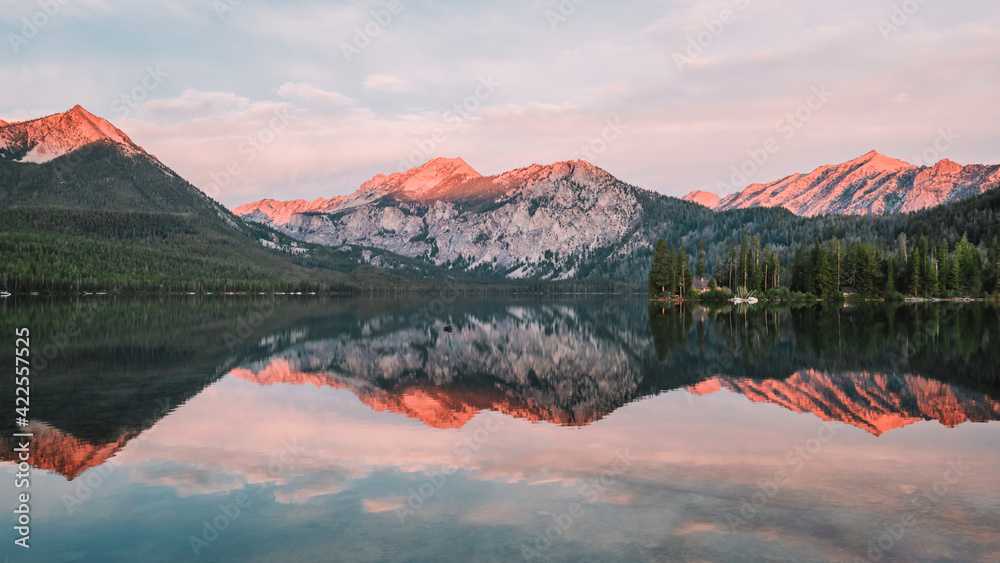 Panoramic landscape of a gorgeous sunrise glow on the peaks of the Sawtooth Mountain Range in the Rocky Mountains and the perfect glass reflections over Pettit Lake in Idaho, USA.