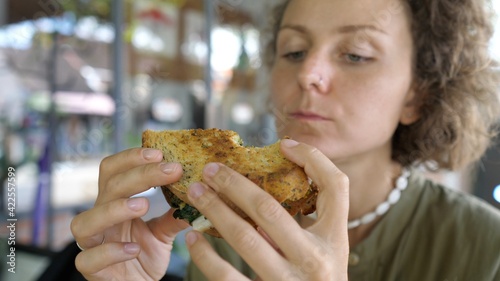 Young caucasian food-loving woman takes a healthy organic vegan sandwich  bites it with appetite and looks at it with appreciation 