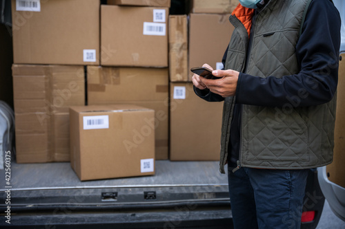 Portrait of a young man shipper with his van during the delivery round in the global pandemic from Covid-19 Coronavirus wearing face mask while checking address with smartphone