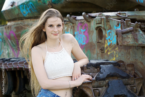 Pretty young woman wears hot pants and top bellyless, leaning against old tank and smiling friendly on sunny summer day in park
