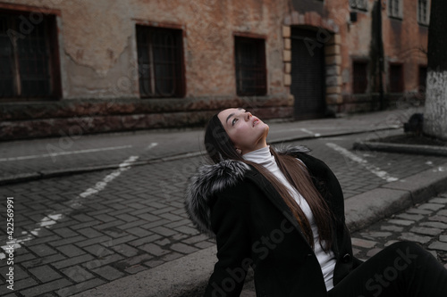 a girl sits on a city street, her head thrown back, looks up