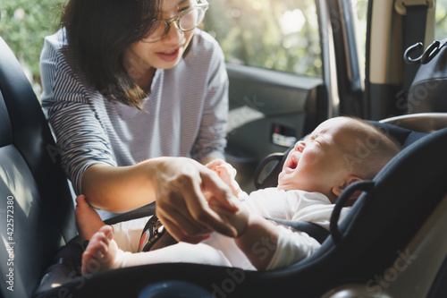 Asian young mother comforting her baby boy crying while putting and fasten seat belts on his car seat in car.
