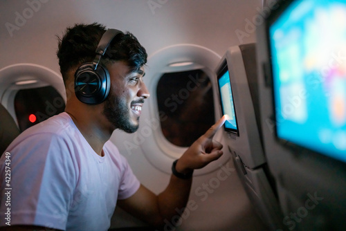 Passenger in airplane touching LCD entertainment screen. Latin american man in plane cabin using smart device listening to music on headphones. photo