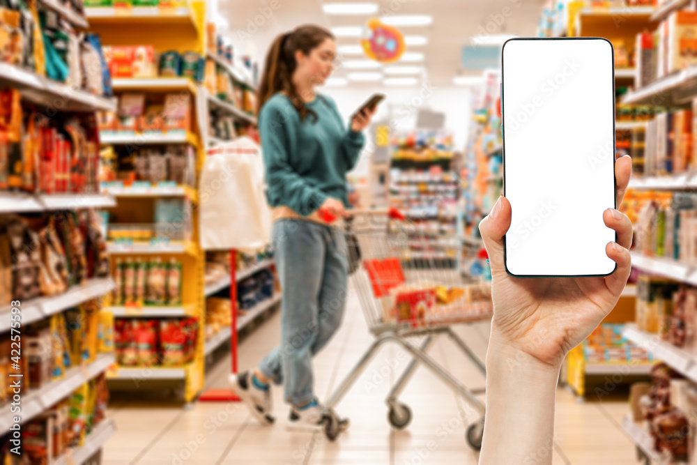 A person's hand holds a mobile phone. Mock up. In the background, a woman using cellphone is pushing a grocery cart in a supermarket, in a blur. The concept of online shopping