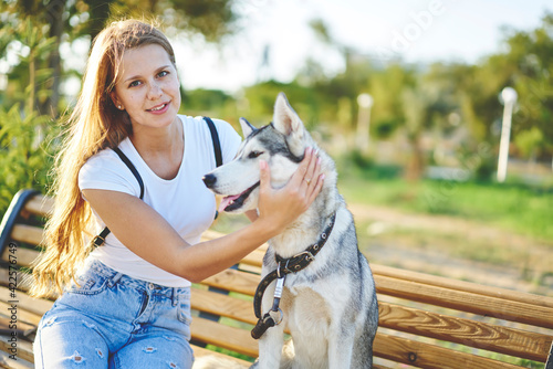 Portrait of young hipster girl with escimo dog resting at park bench looking at camera during walk daytime, teenage female owner sitting near siberian husky enjoying leisure during summer sunny day