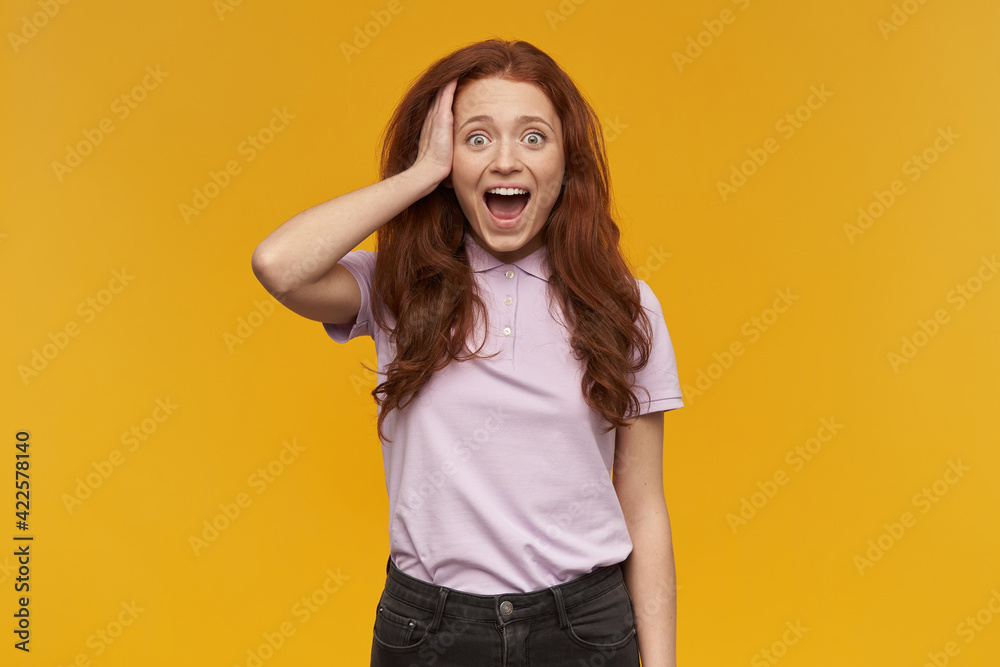 Fototapeta premium Shocked, positive woman with long ginger hair. Wearing pink t-shirt. People and emotion concept. Touching her head, surprised. Watching at the camera, isolated over orange background