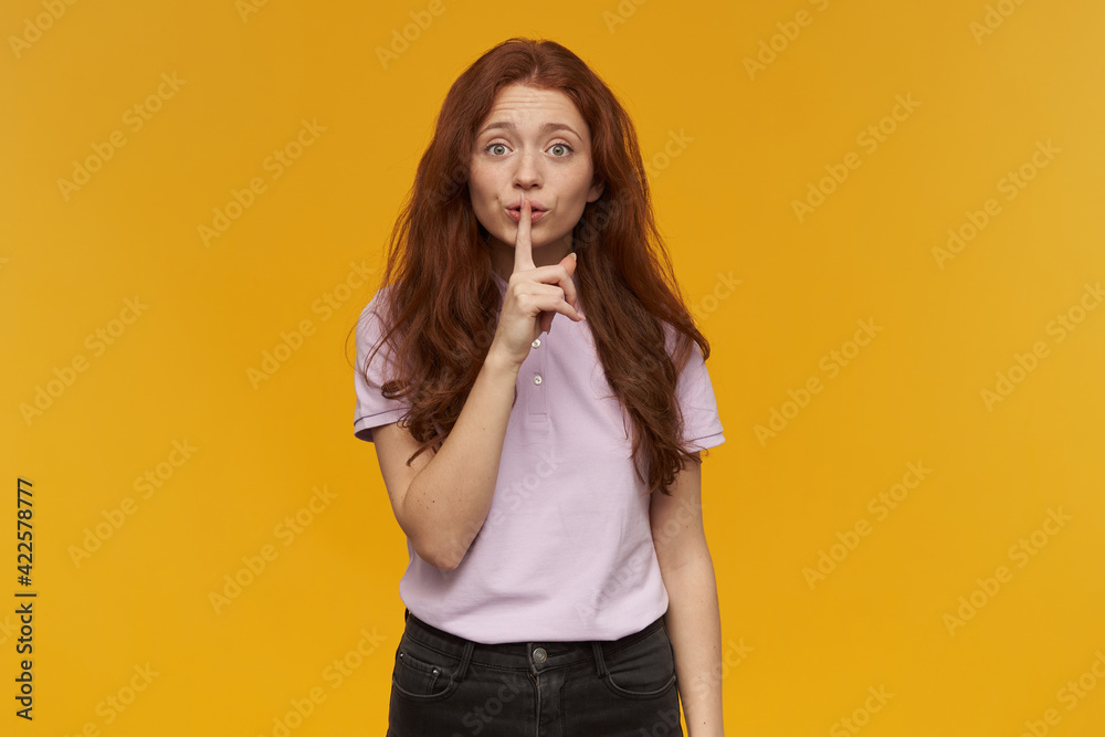 Cute, attractive woman with long ginger hair. Wearing pink t-shirt. People and emotion concept. Showing silence sign, asks to be quiet. Watching at the camera, isolated over orange background Cute, attractive woman with long ginger hair. Wearing pink t-shirt. People and emotion concept. Showing silence sign, asks to be quiet. Watching at the camera, isolated over orange background