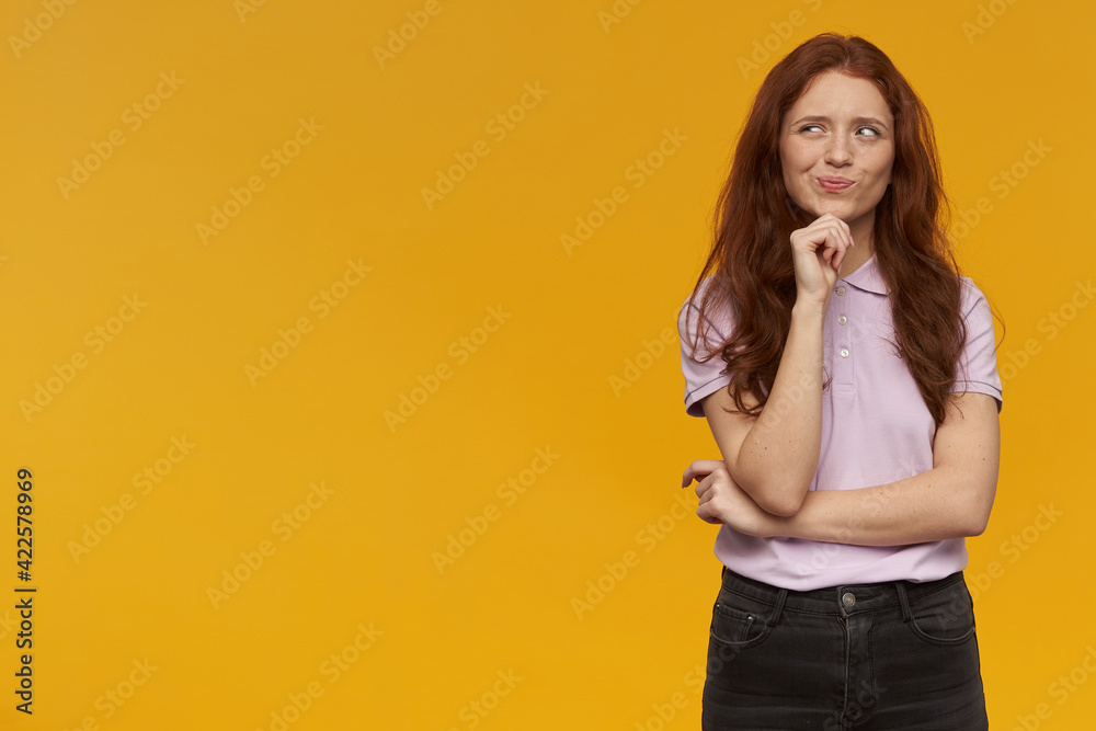 Wonder looking girl, attractive redhead woman with long hair. Wearing pink t-shirt. Emotion concept. Touching her chin and dreaming. Watching to the left at copy space, isolated over orange background Wonder looking girl, attractive redhead woman with long hair. Wearing pink t-shirt. Emotion concept. Touching her chin and dreaming. Watching to the left at copy space, isolated over orange background