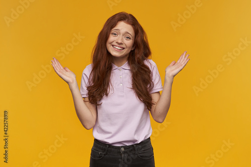 Unsure looking girl, attractive redhead woman with long hair. Wearing pink t-shirt. Emotion concept. Shrugs her shoulders and broadly smiling. Watching at the camera, isolated over orange background
